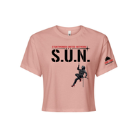 Stuntwomen United light pink women’s cropped T-shirt shown from the front, featuring S.U.N. logo with rappelling stuntwoman graphic in red heels and Stunt Team sleeve logo.