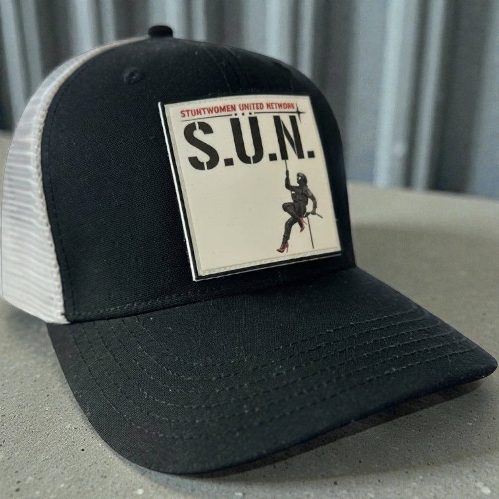 Close-up of a black and white Stuntwomen United Network Richardson trucker hat featuring a velcro-backed rubber patch on the front panel.